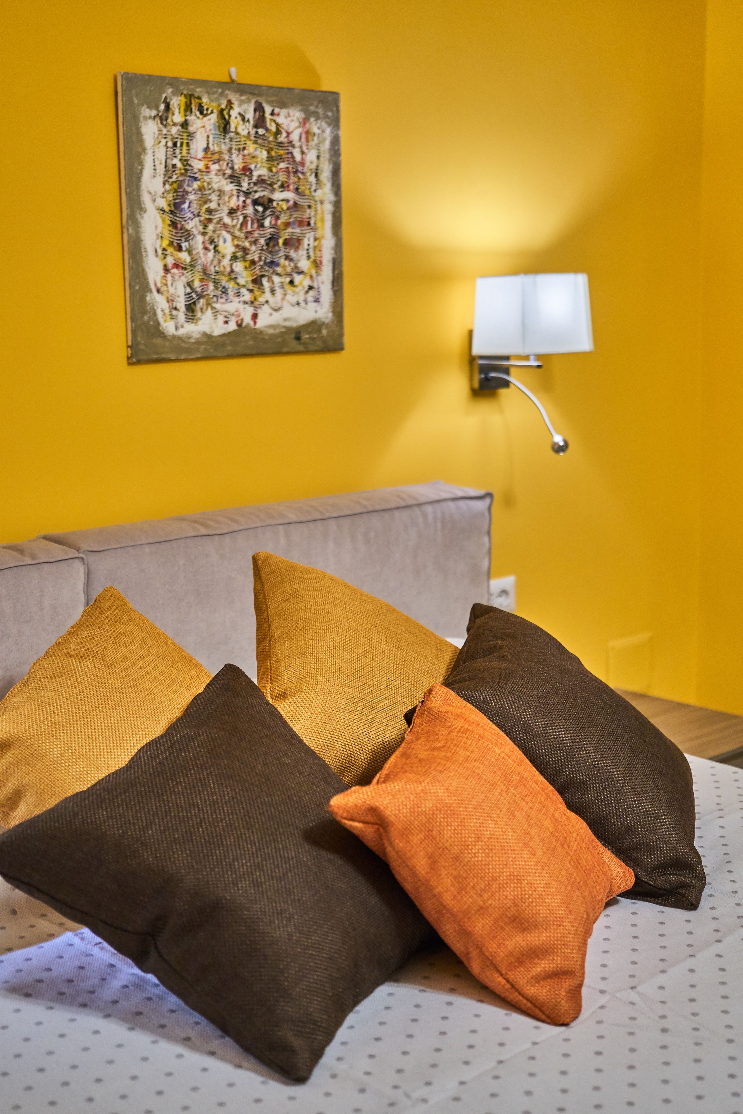 A bed with multiple pillows and a wall-mounted lamp in a room with a yellow wall.