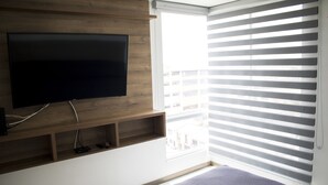A modern bedroom with a large flat-screen TV mounted on a wooden wall, a window with blinds, and a bed with a gray quilt. - Apartamento Granate (Pereira)