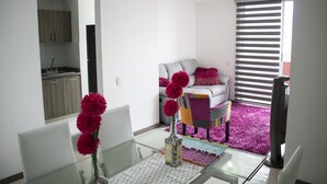 A modern living room with a glass dining table, a sofa with colorful cushions, and a kitchen area visible through an open door. - Apartamento Granate (Pereira)