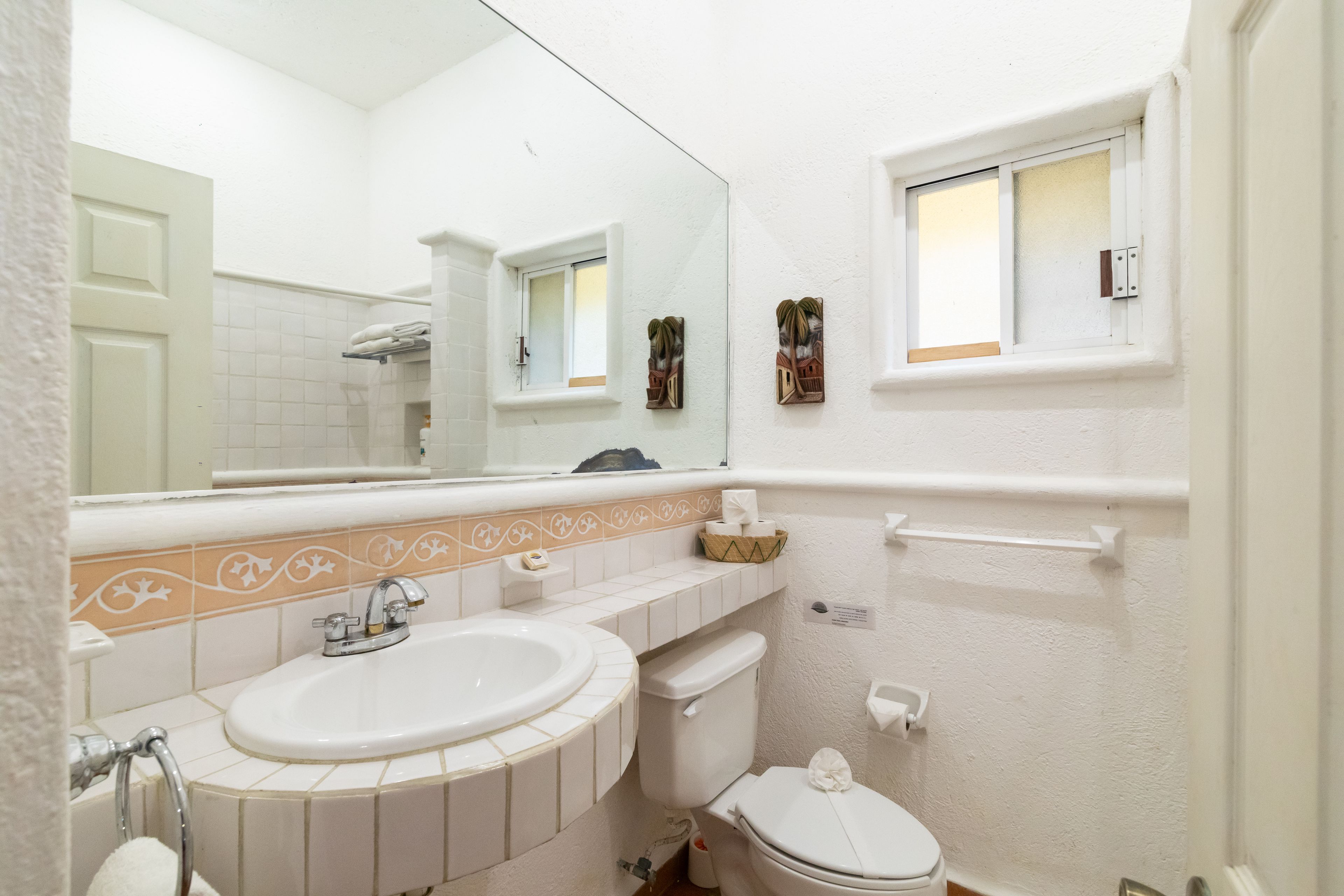 A bathroom with a white sink, toilet, and mirror. There is a window with a view of palm trees and a shelf with toiletries.