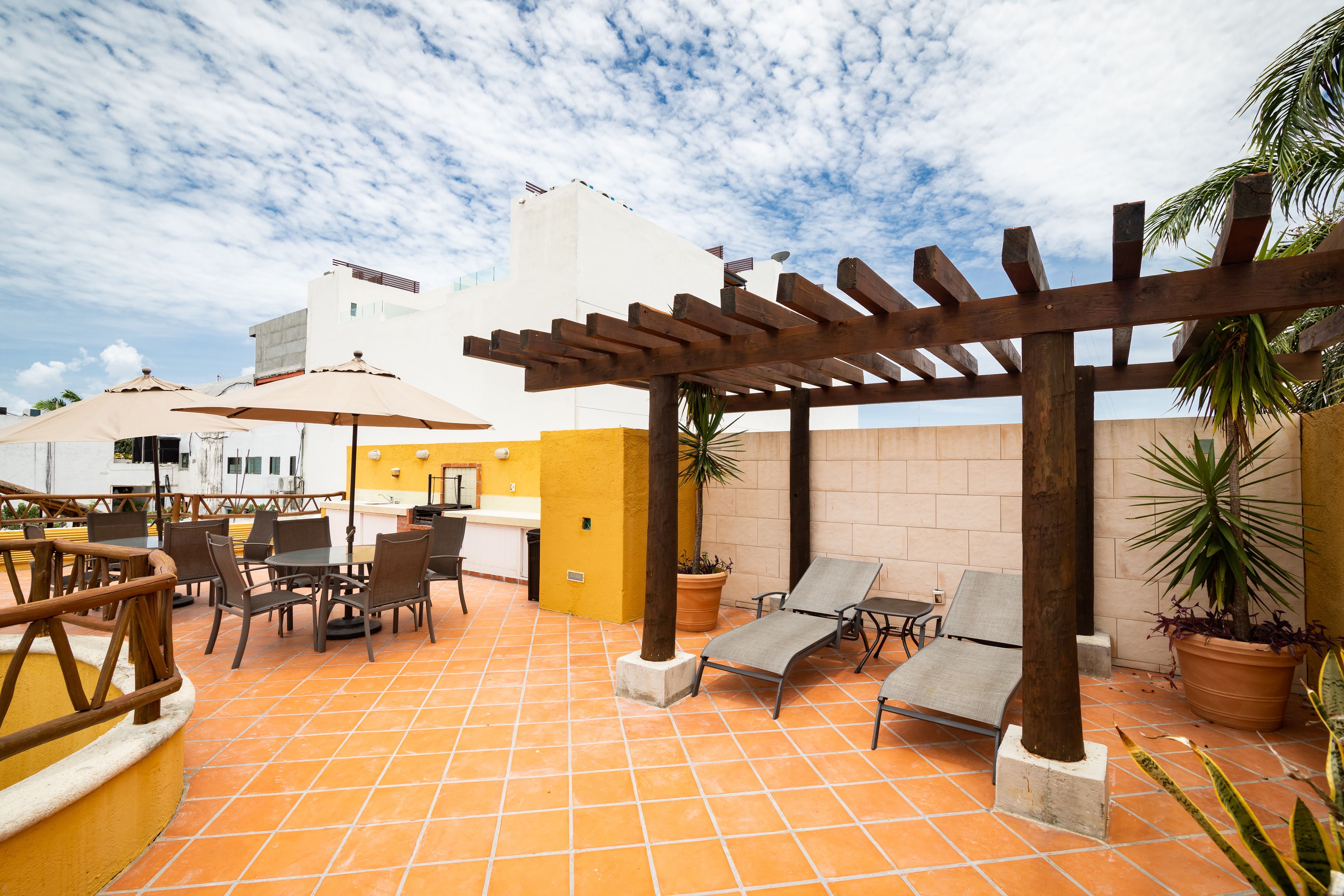 An outdoor patio with tiled flooring, wooden pergola, lounge chairs, and potted plants.
