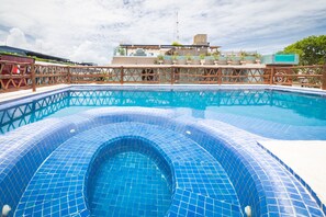 A swimming pool with blue mosaic tiles, surrounded by a wooden deck and a view of a building with a satellite dish.