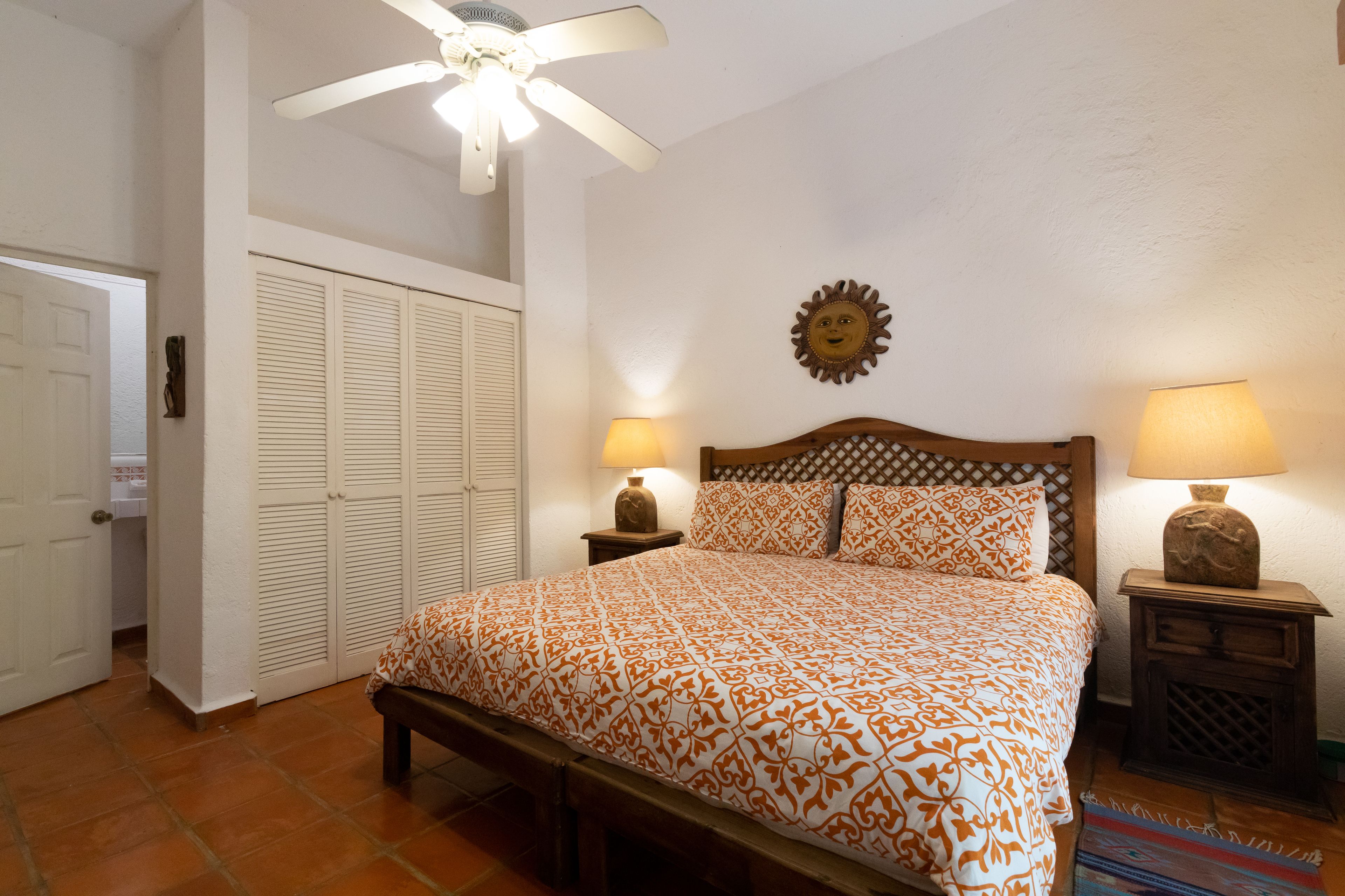 Une chambre à coucher comprenant un lit, deux lampes de chevet, un ventilateur de plafond, une décoration murale en forme de soleil et une porte donnant sur une salle de bain.