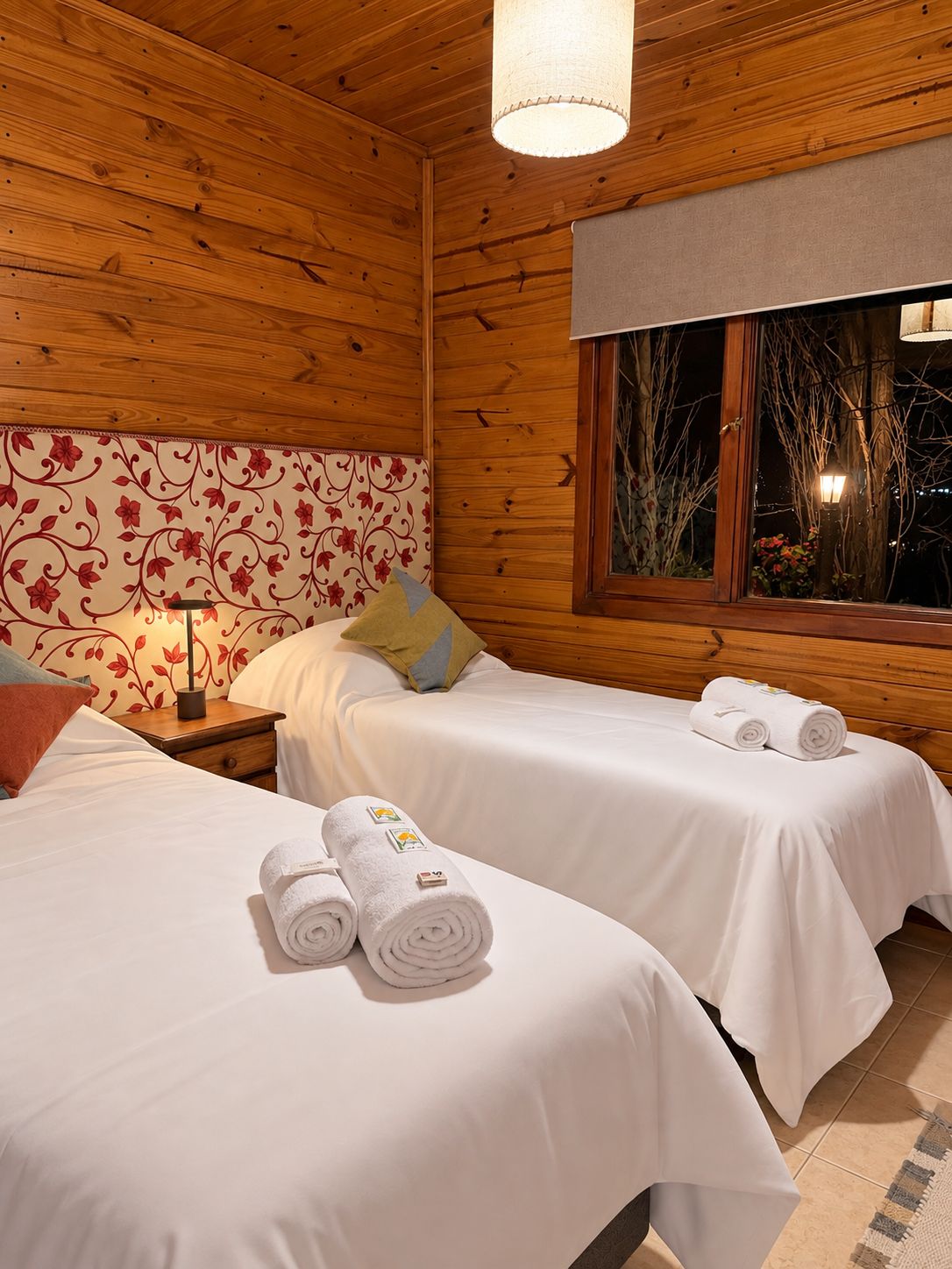 A wooden cabin room with two beds, a nightstand, a lamp, and a window with a view of bare trees.