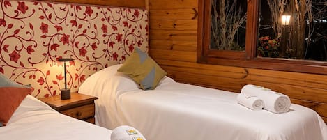 Une chambre dans un chalet en bois, avec deux lits, une table de chevet, une lampe et une fenêtre donnant sur des arbres sans feuilles.