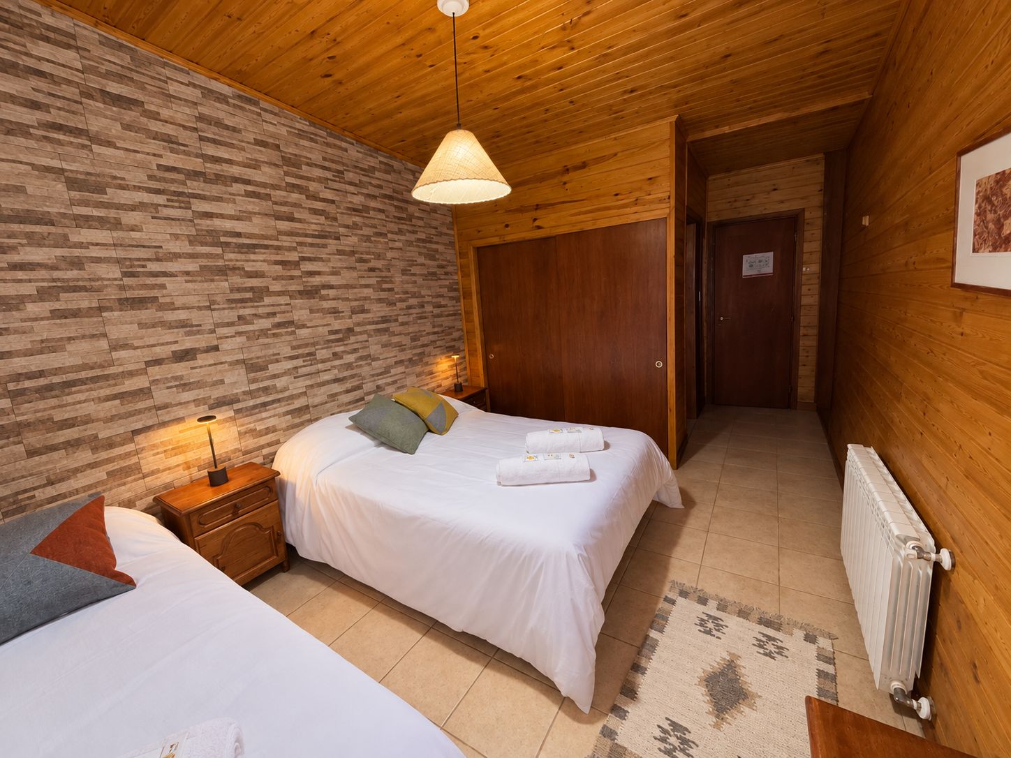A hotel room with two beds, a wooden ceiling, a brick-patterned wall, a wooden door, and a radiator.