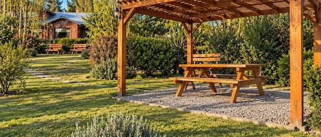 Una pérgola de madera con una mesa de picnic, rodeada por un jardín cuidado con césped y un sendero de grava.