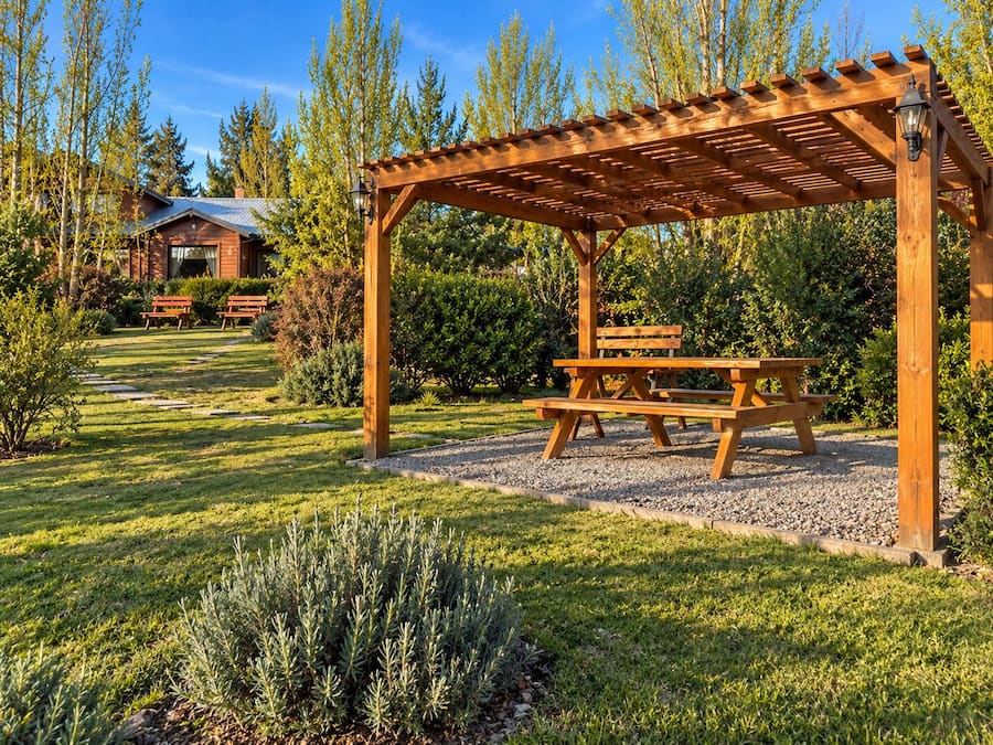 Una pérgola de madera con una mesa de picnic, rodeada por un jardín cuidado con césped y un sendero de grava.