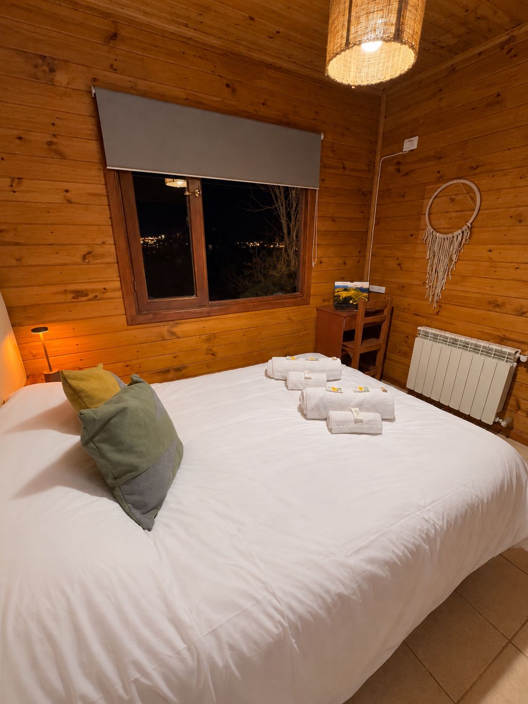 A wooden room with a bed, a window, a wall-mounted dreamcatcher, and a radiator.