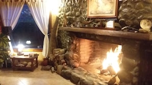 Une cheminée en pierre avec un feu, une étagère en bois rustique au-dessus, et une fenêtre avec des rideaux.