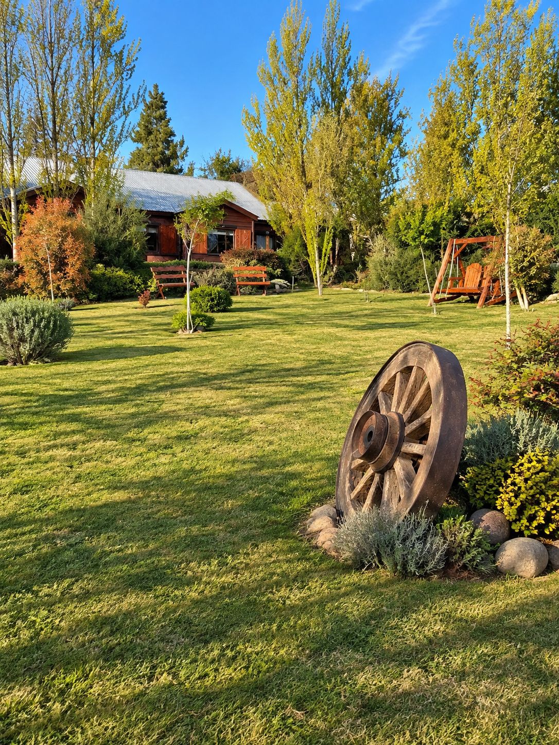 Uma casa rústica com uma grande roda em primeiro plano, rodeada por um jardim bem cuidado e árvores.