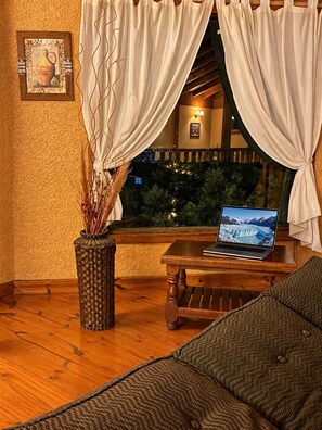 A cozy room with a wooden floor, a wicker chair, a small table with a laptop, and a view of a snowy landscape through a window with white curtains. - Hosteria Rukahue (El Calafate)