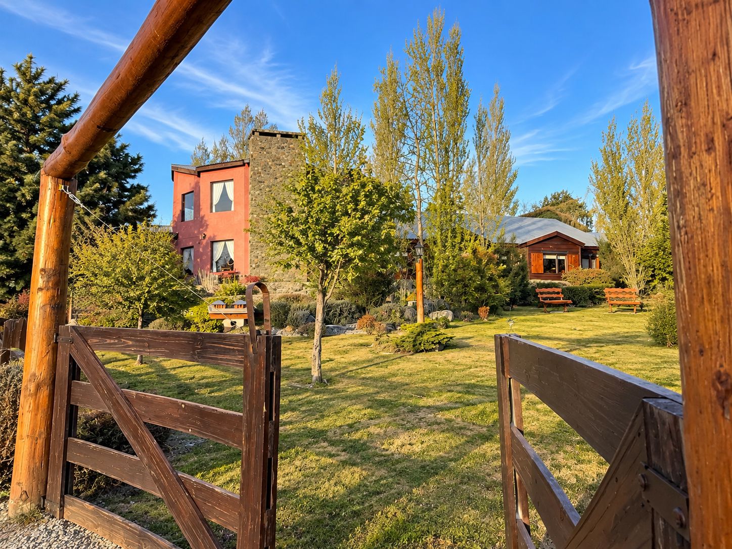 Una verja de madera da paso a un área con césped, con un edificio rojo y una cabaña más pequeña al fondo.