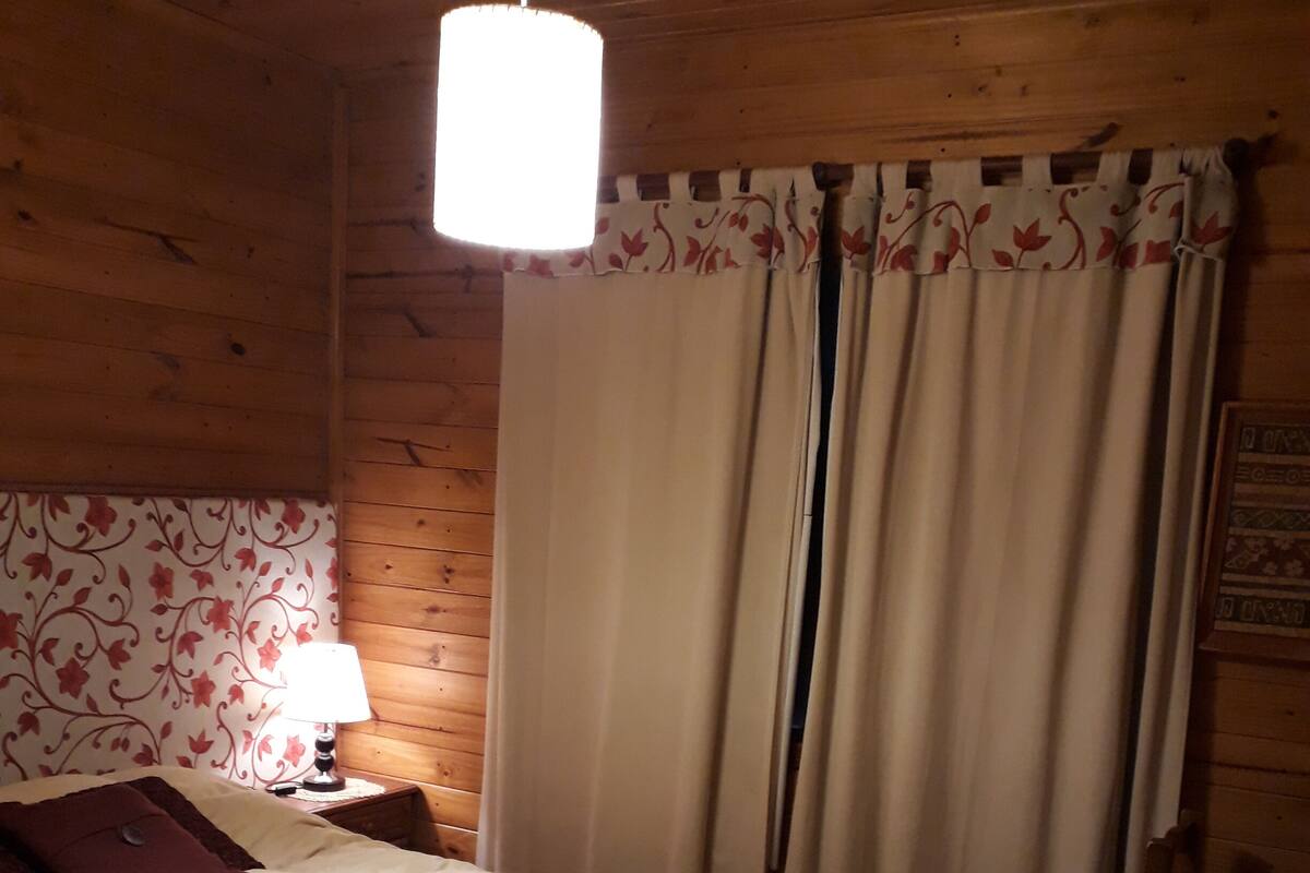 A wooden cabin bedroom with a bed, a nightstand, a lamp, and a window with curtains.