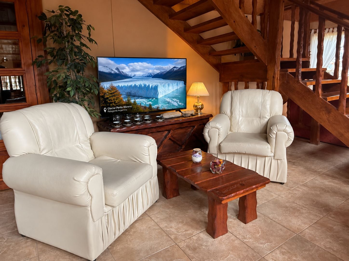 A living room with two white leather sofas, a wooden coffee table, a television displaying a glacier, and a staircase.