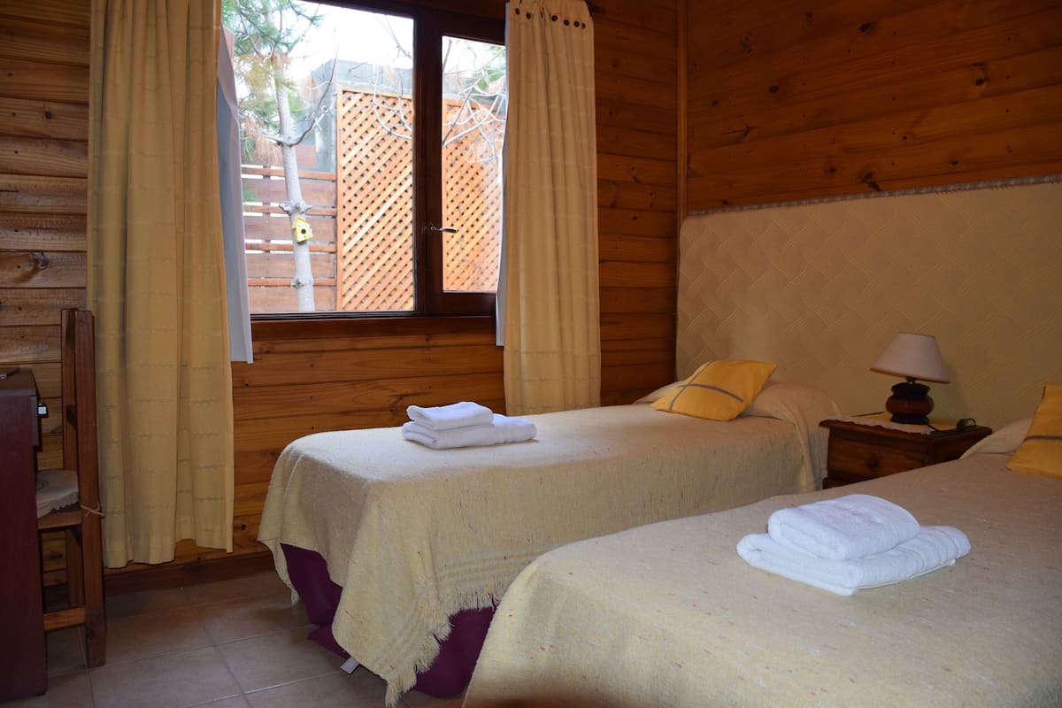 A wooden cabin room with two beds, a window with curtains, a nightstand with a lamp, and a wooden chair.