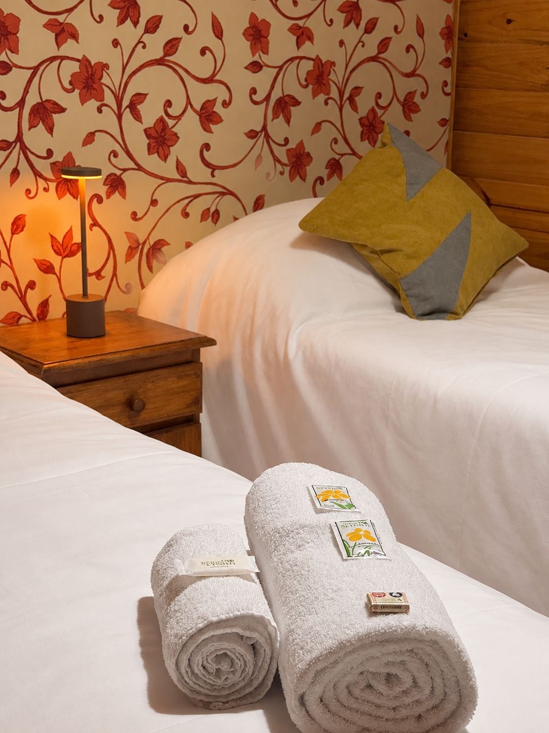 A neatly made bed with white bedding, a wooden nightstand with a lamp, a floral wallpaper, and two rolled towels with hotel branding.