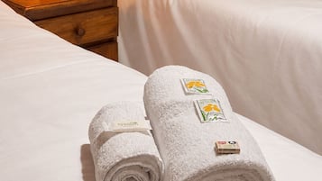 A neatly made bed with white bedding, a wooden nightstand with a lamp, a floral wallpaper, and two rolled towels with hotel branding.
