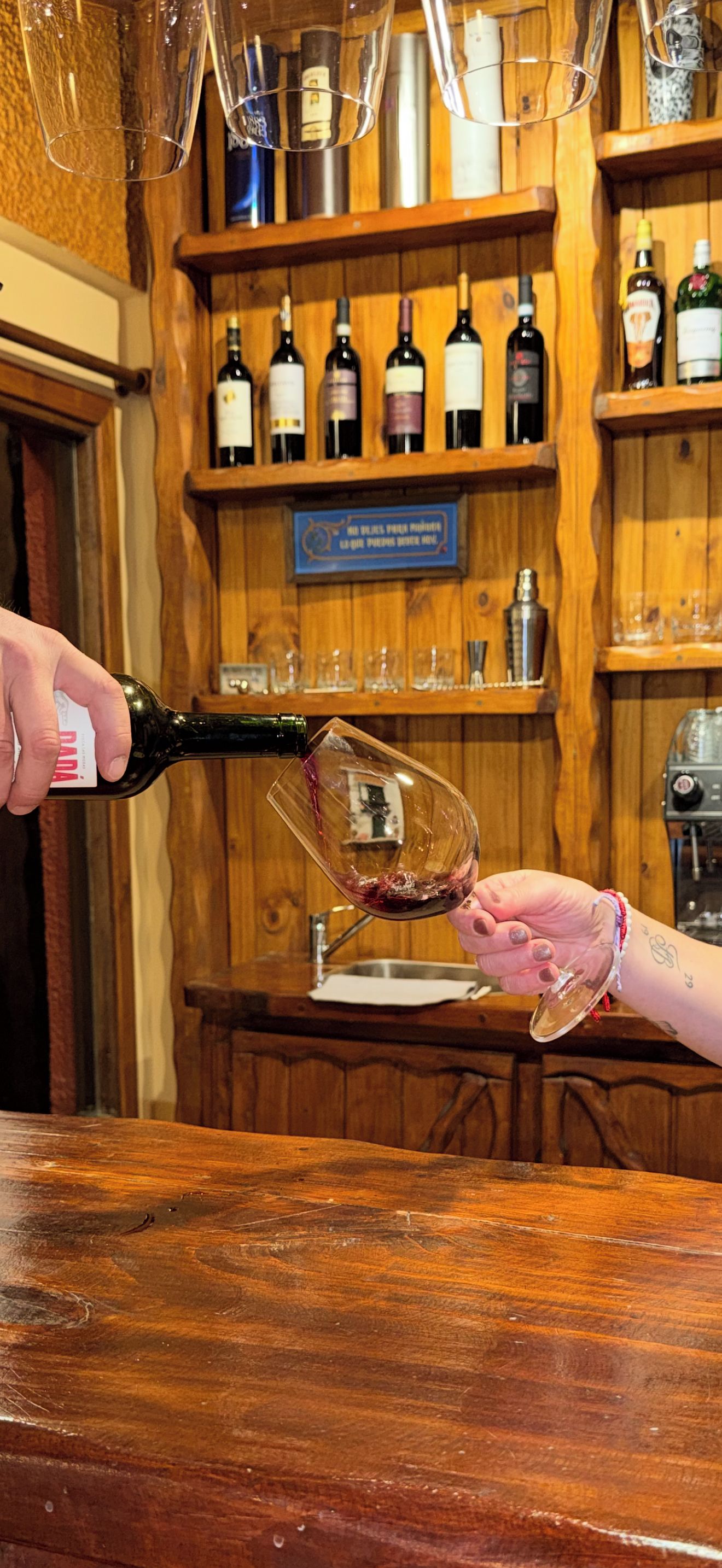 Eine Person gießt Wein in ein Glas an einer Bar mit Holzböden und verschiedenen Flaschen.