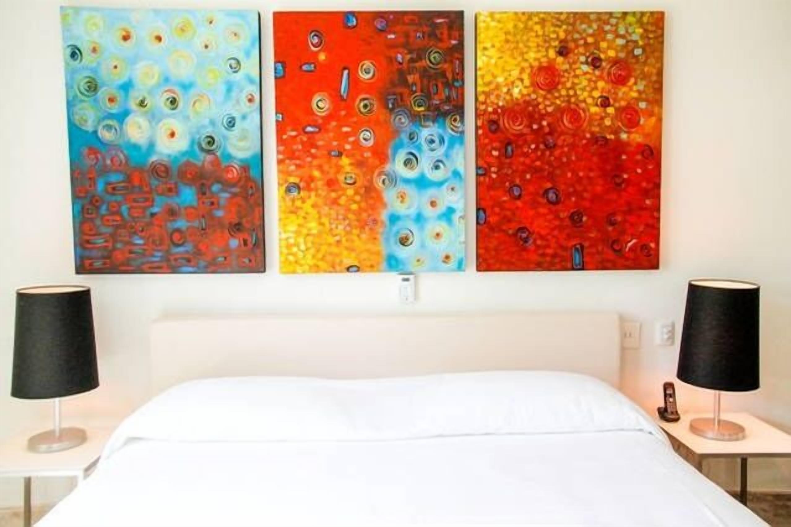 A hotel room with a bed, two bedside tables, and three colorful abstract paintings on the wall.