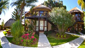 Un resort con una estructura de techo de paja, un área de estar al aire libre techada y un jardín bien cuidado con flores rosas.