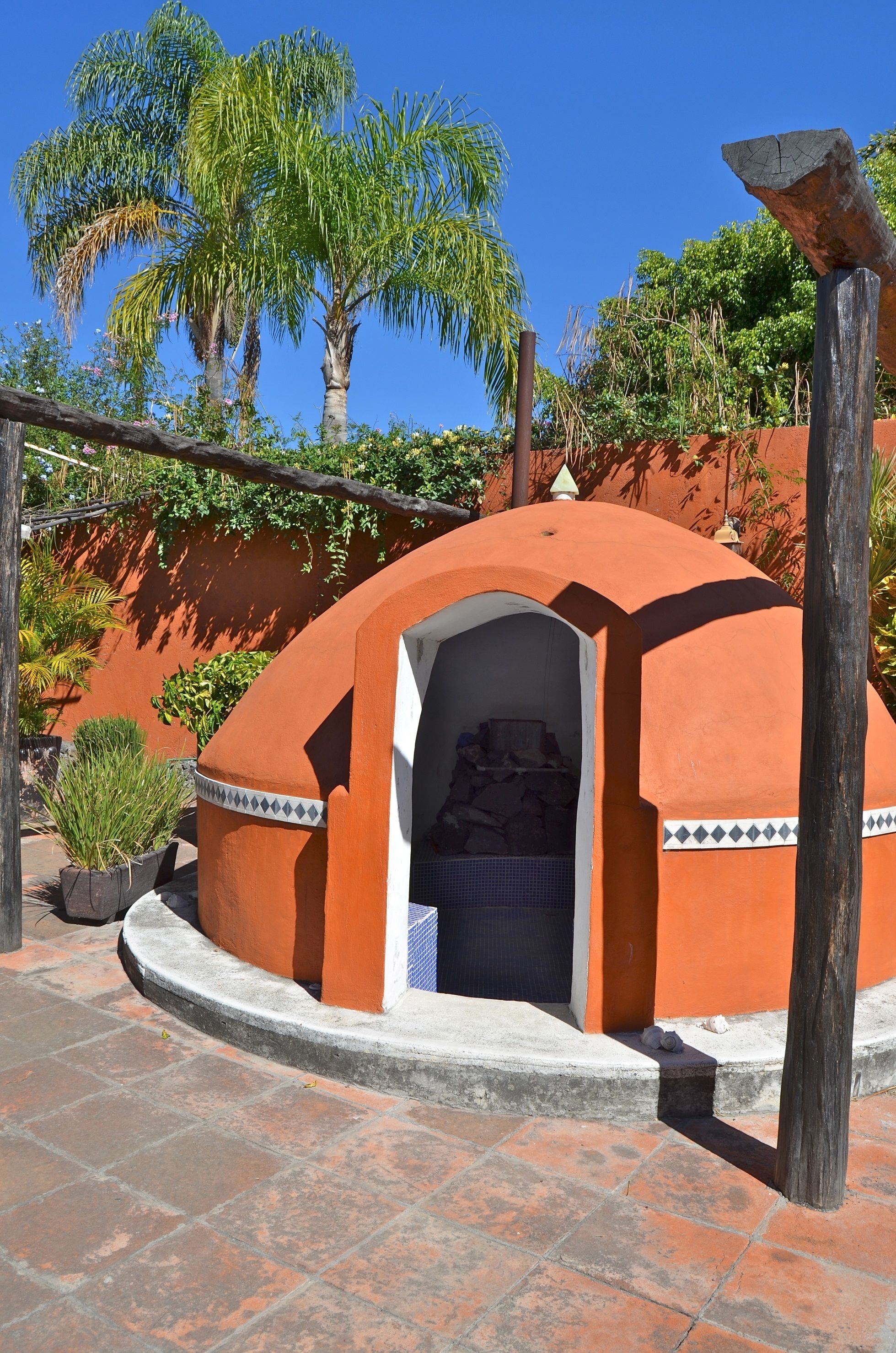A small, dome-shaped structure with an open doorway, surrounded by palm trees and a red wall.