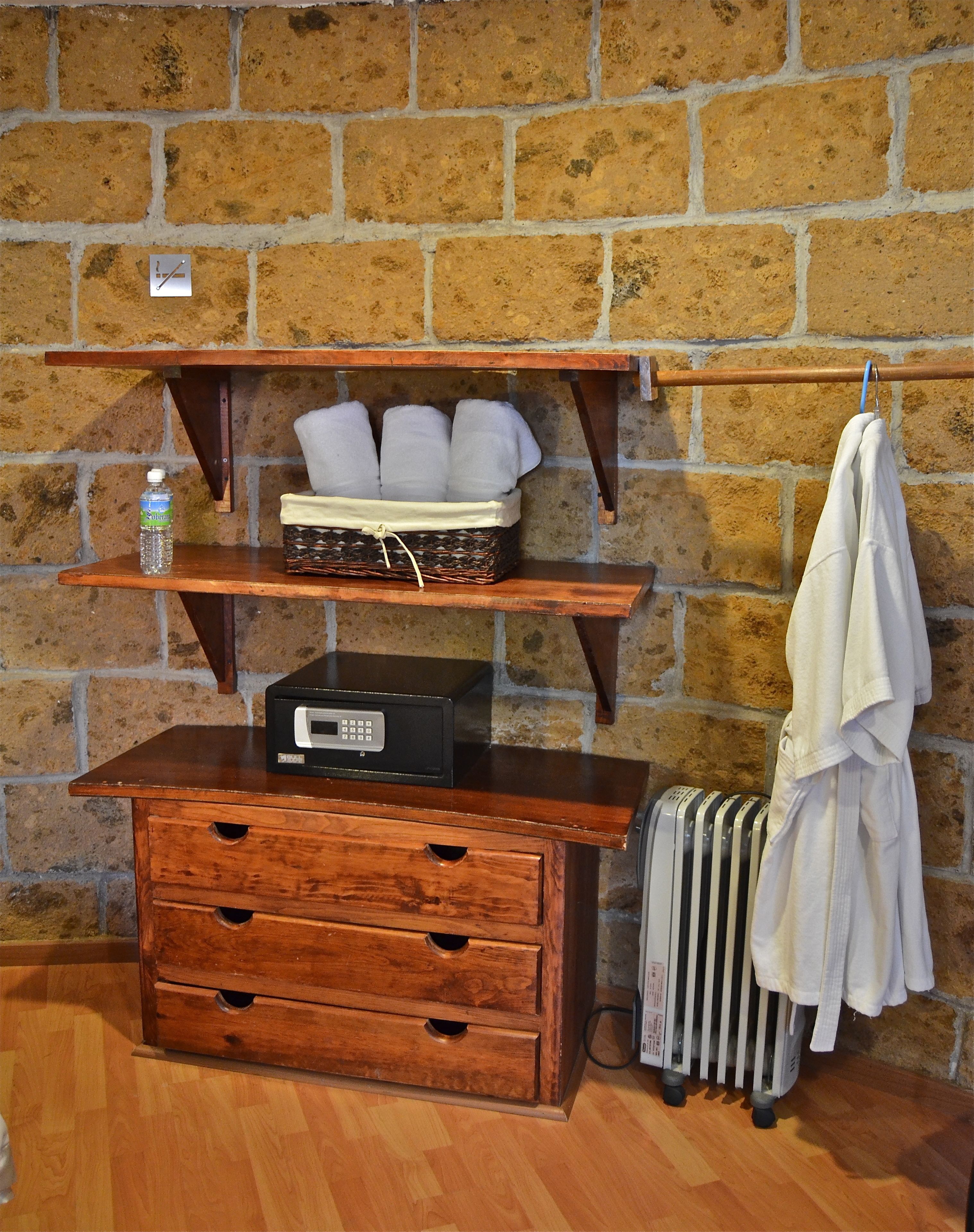 A wooden chest of drawers, a microwave, a basket with towels, a radiator, and a hanging towel rack against a brick wall.