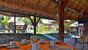 Un área de comedor al aire libre con mesas dispuestas para dos, sombrillas de paja y vistas a un jardín ajardinado.