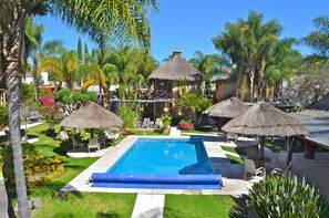 Piscina rodeada de palmeras y construcciones con techo de paja.