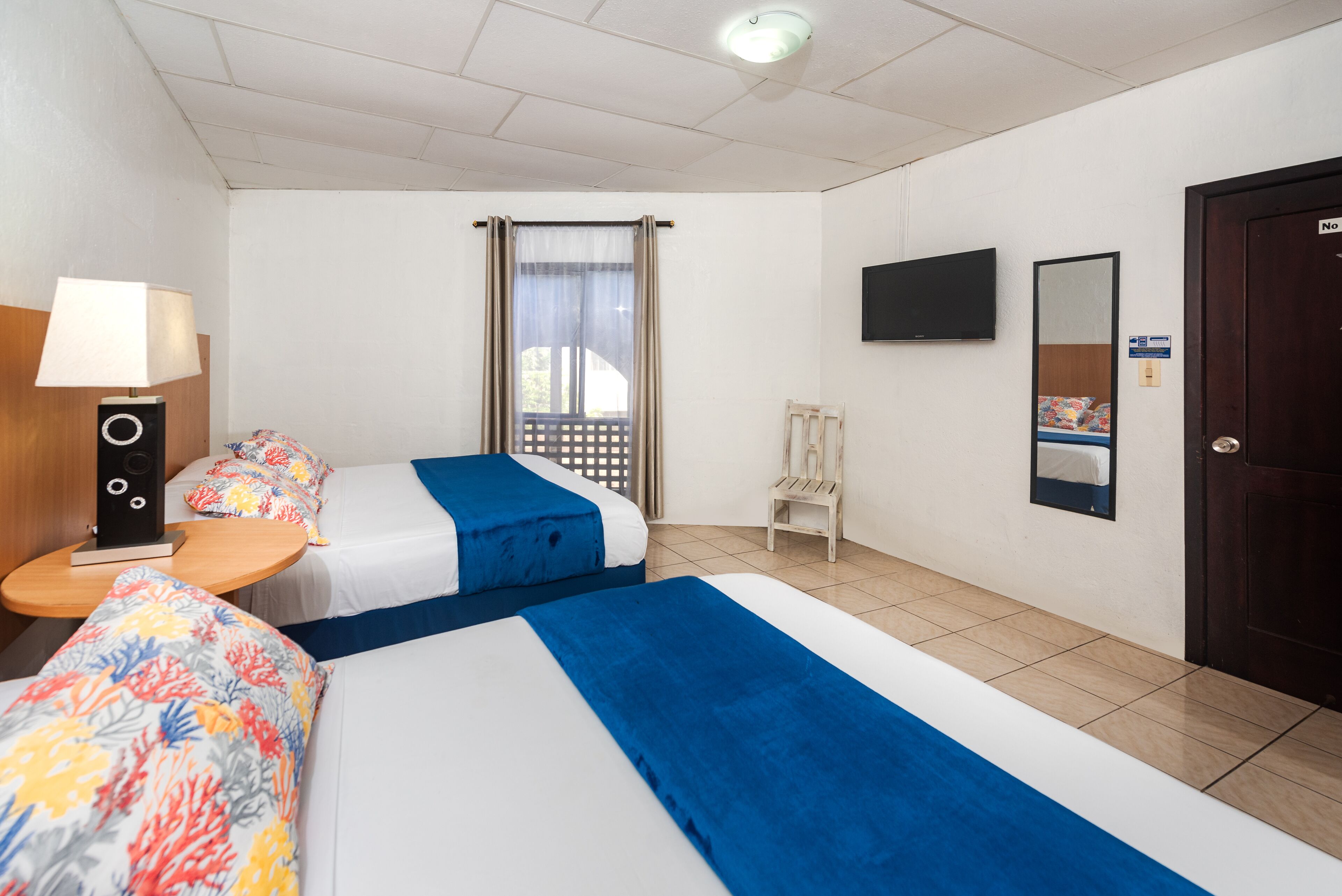 A hotel room with two beds, a TV, a mirror, and a window with curtains.