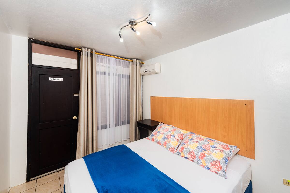 Ein Schlafzimmer mit Bett, Nachttisch, Holz-Kopfteil, einer Tür und einem Fenster mit Vorhängen.