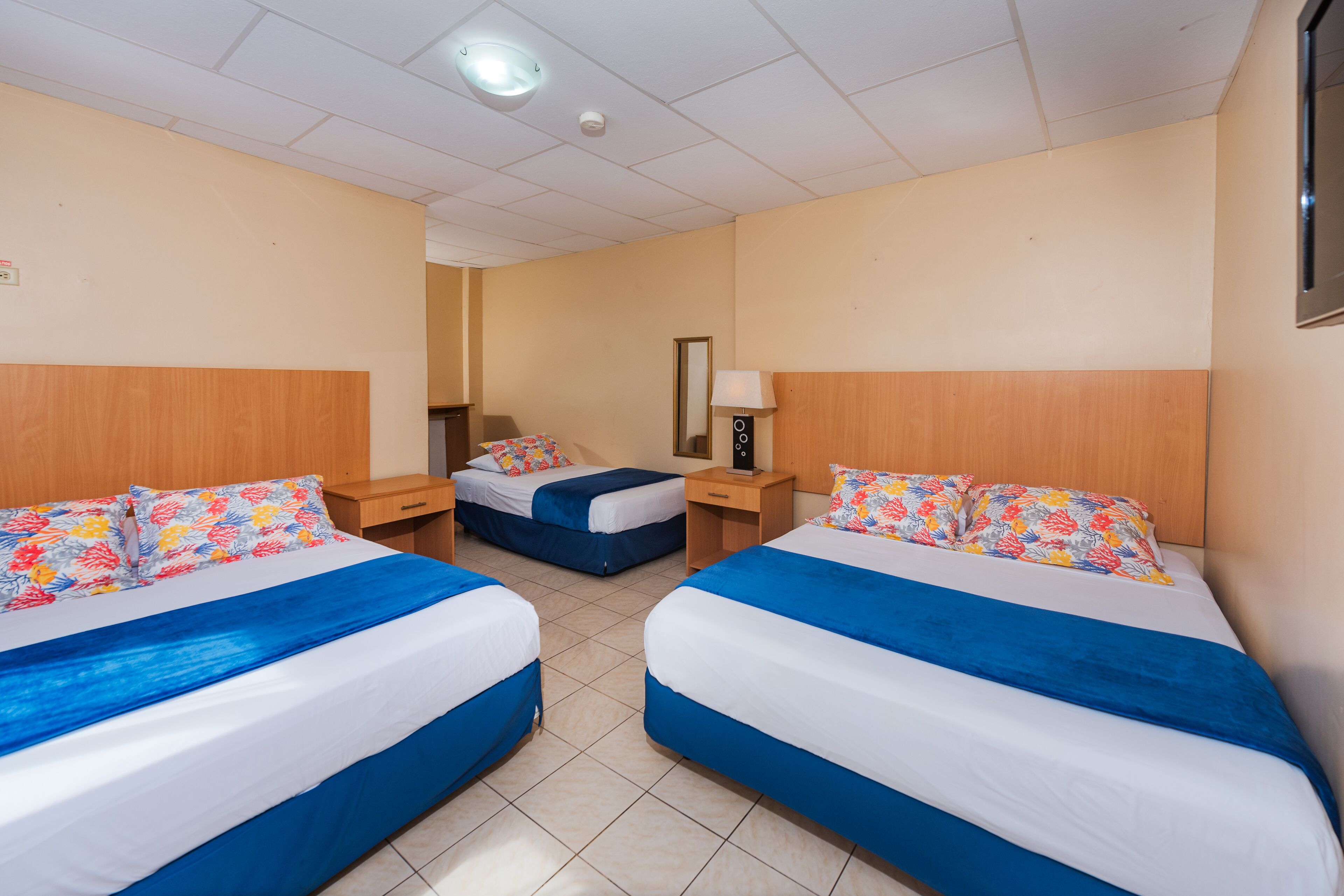 A hotel room with two beds, each with blue and white bedding and floral-patterned pillows. There is a nightstand with a lamp and a small table between the beds. The room has beige walls and a tiled floor.
