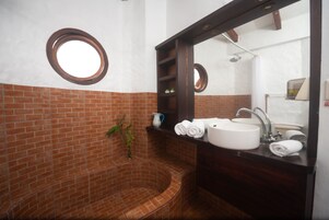 A bathroom with a brick wall, a round window, a sink, a mirror, and a shower.