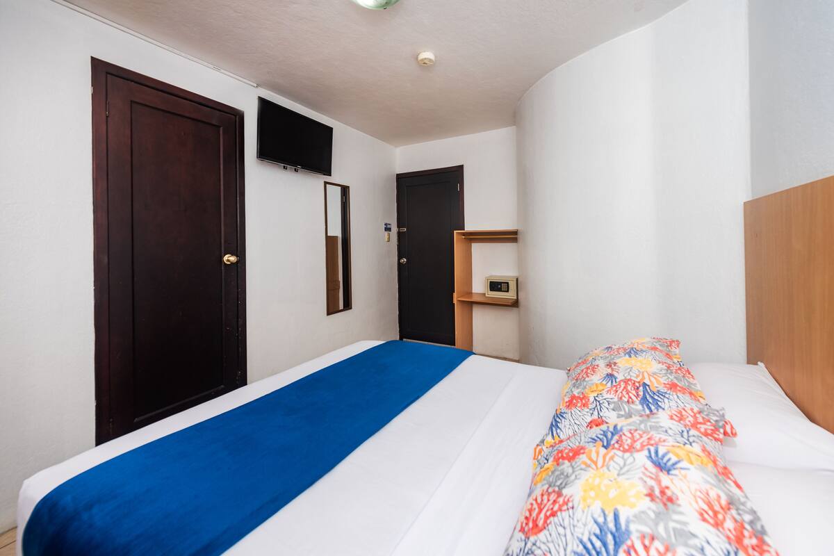 Ein Hotelzimmer mit Bett, einem an der Wand befestigten Fernseher, einer Holztür und einem kleinen Regal mit einer Uhr.