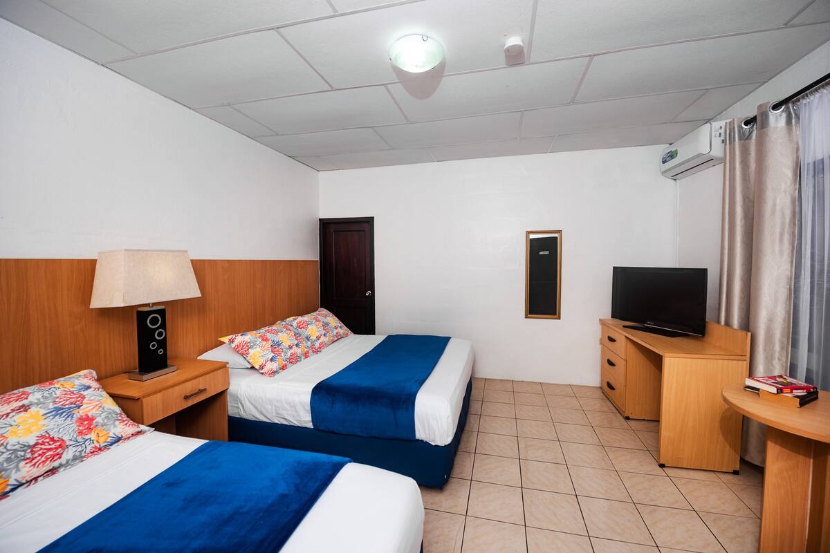 Ein Hotelzimmer mit zwei Betten, einem Schreibtisch, einem Fernseher und einer Klimaanlage.