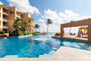 A swimming pool with a view of the ocean, palm trees, and a building with balconies. - Condo El Faro Coral By Playa Paradise (Playa del Carmen)