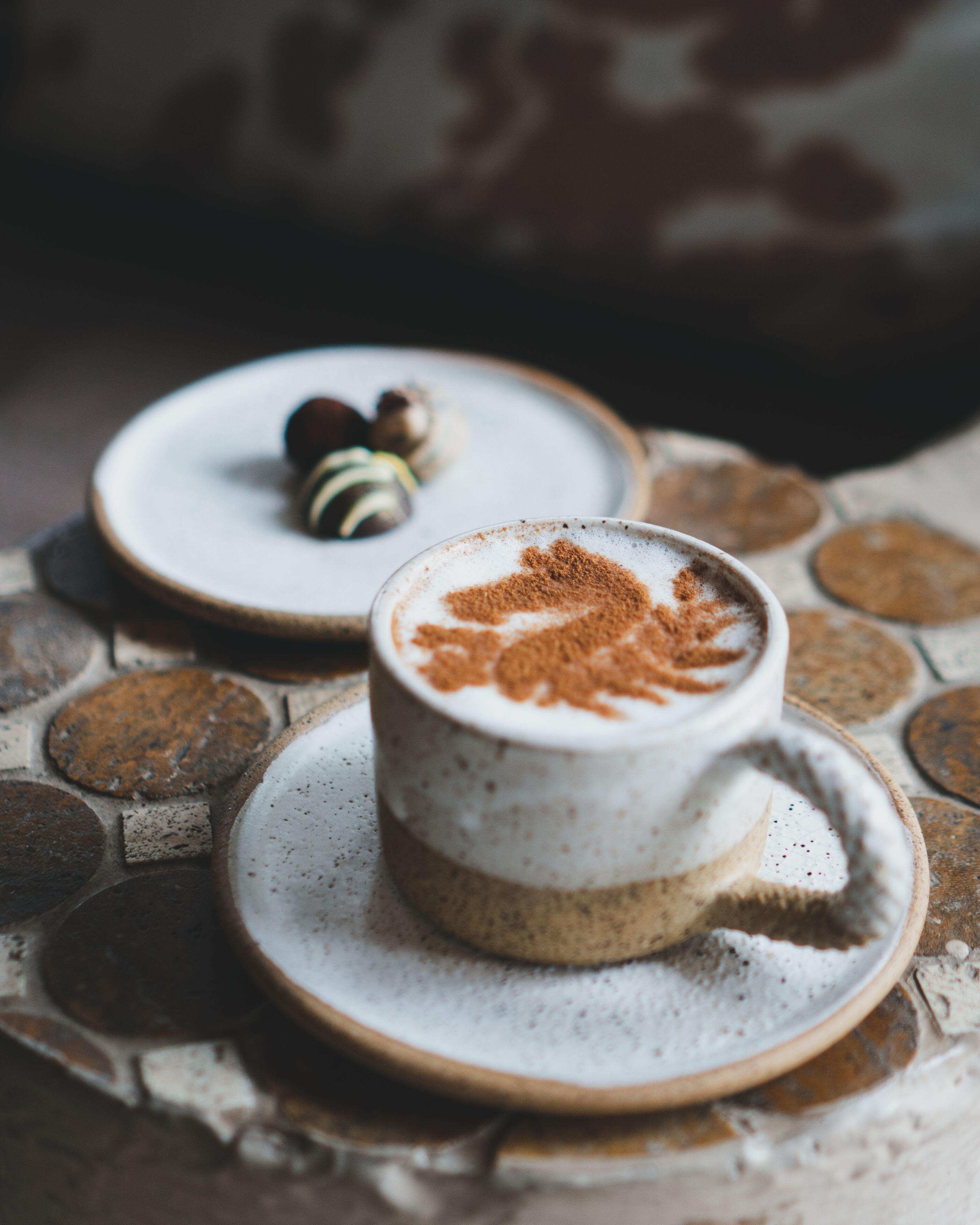A cup of coffee with a latte art design on a saucer, placed on a table with a rustic, circular tile pattern.