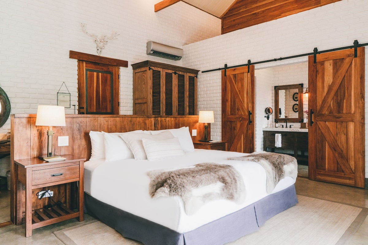 Un dormitorio con una cama grande, cabecera de madera, dos mesitas de noche con lámparas, un aparador y una puerta corredera de estilo rústico.