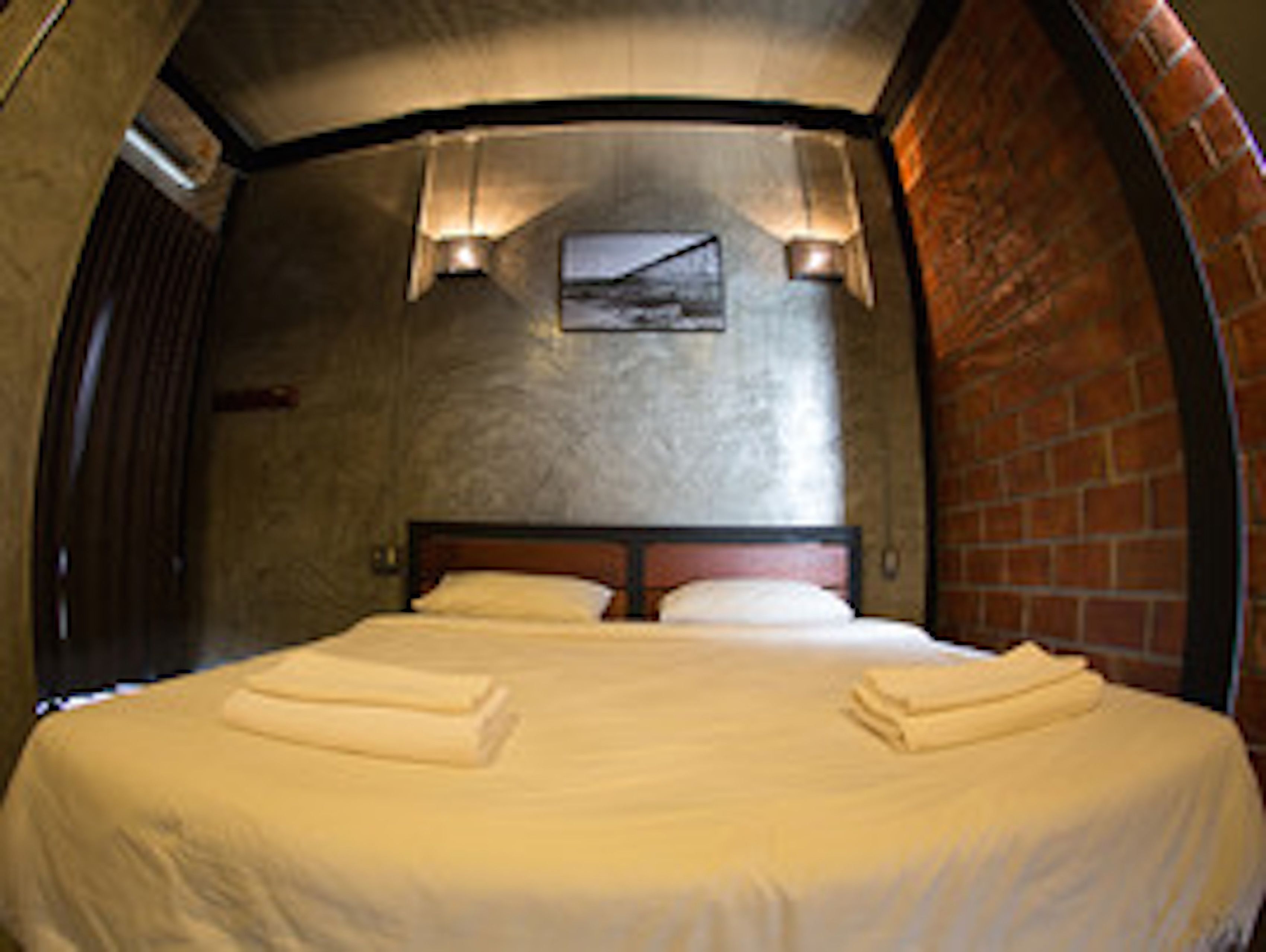 A room with a bed, a headboard, a wall-mounted picture, and a brick wall.