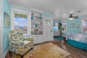 Een kamer met een stoel in strandthema, een wit inbouwkast, een bed met een blauw dekbed en een plafondventilator.