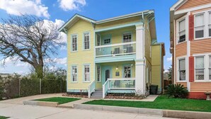 A two-story house with a front porch, a small front yard, and a tree in the background. - BEAUTIFUL 5-BEDROOM HOUSE WITH HEATED SWIMMING POOL, GAS GRILL, LARGE DECK (Galveston)