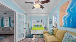 A room with a bed, a sofa, a ceiling fan, and a large painting. - BEAUTIFUL 5-BEDROOM HOUSE WITH HEATED SWIMMING POOL, GAS GRILL, LARGE DECK (Galveston)