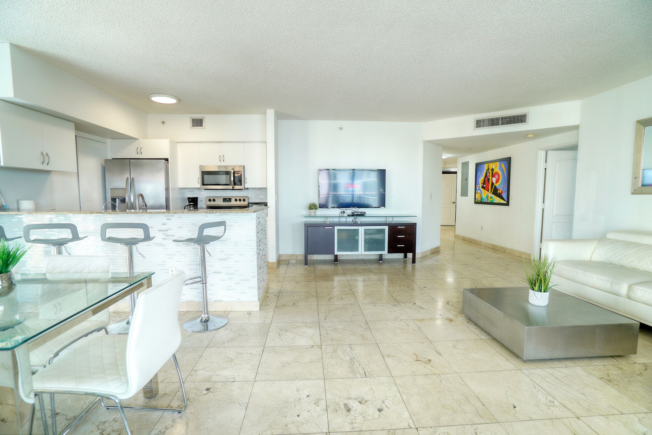 Eine moderne Küche mit Kochinsel, ein Essbereich mit Glastisch und Stühlen sowie ein Wohnzimmer mit Sofa und Wand-Fernseher.