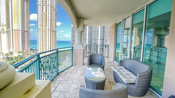 A balcony with wicker furniture, a glass table, and a view of tall buildings and the ocean.