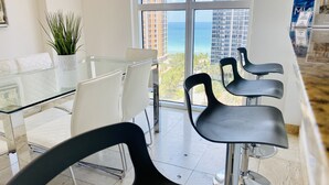 A modern hotel lobby with a glass table and chairs, a potted plant, and a view of tall buildings and the ocean.