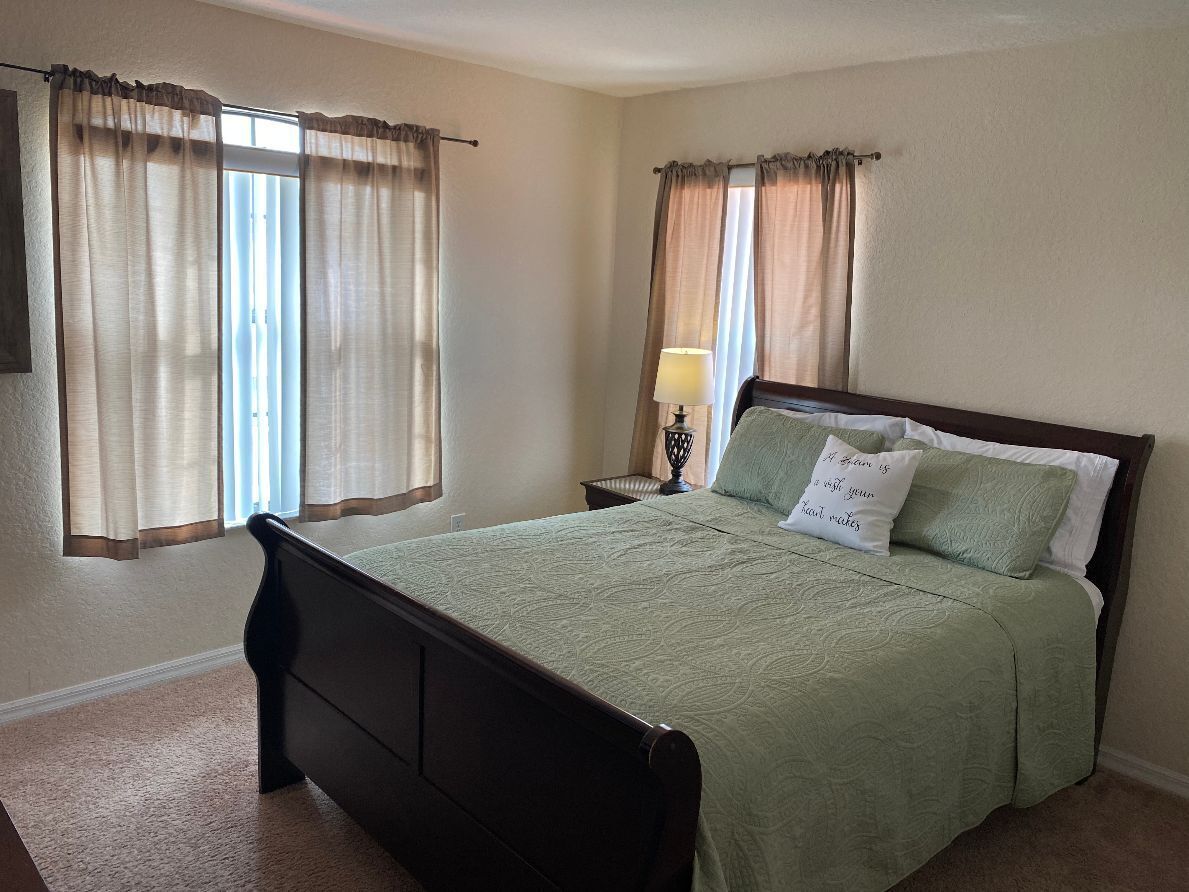 A neatly made bed with a green bedspread and two pillows, a bedside lamp, and curtains on a window.