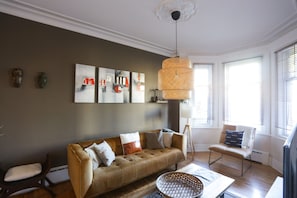 A living room with a brown sofa, a coffee table, and a wicker lamp. There are abstract paintings on the wall and a window with blinds.