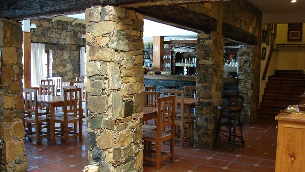Un interior rústico con pilares de piedra, mesas y sillas de madera. Se observa un área de bar con botellas y vasos al fondo.