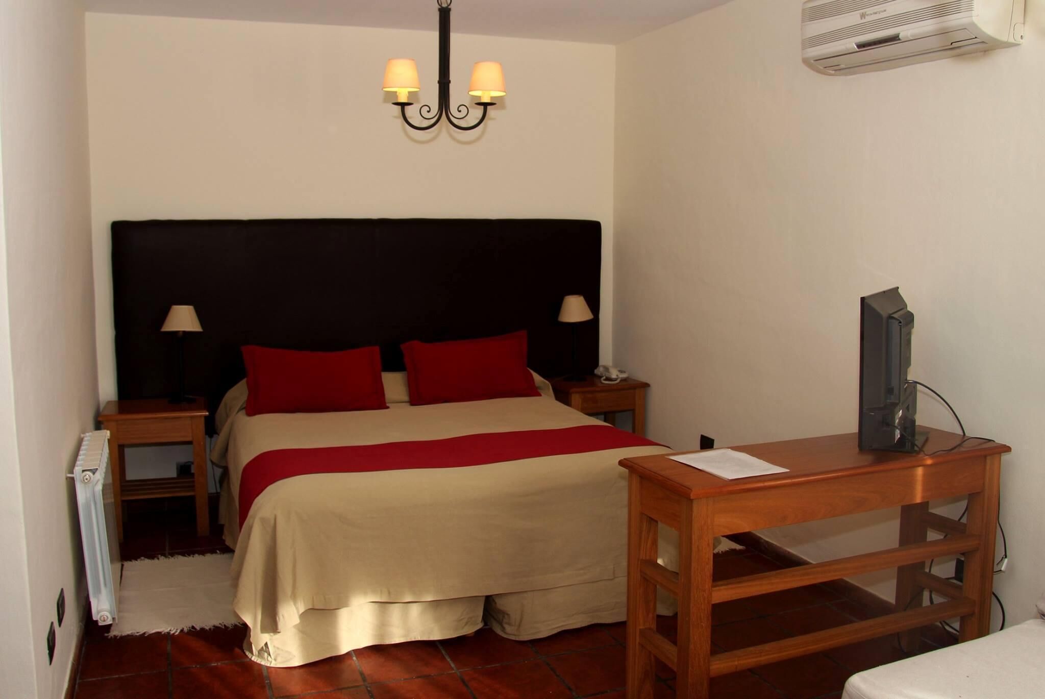 A bedroom with a bed, two bedside lamps, a TV on a wooden table, and an air conditioner on the wall.
