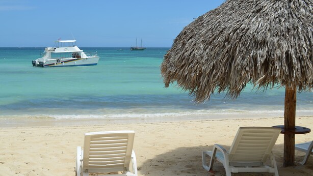 Een strand met twee witte ligstoelen, een rieten parasol en een boot in het water.